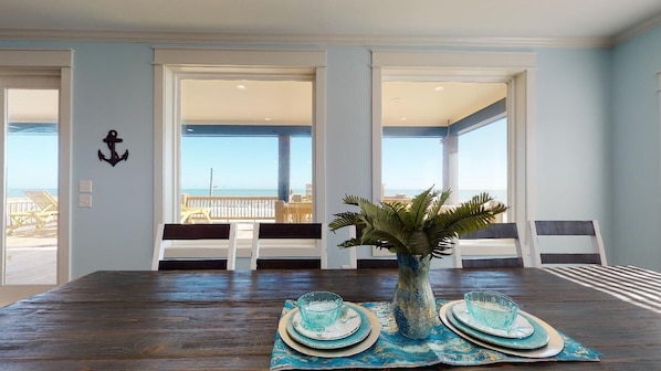 A dining area with a wooden table, a vase with a plant, and a view of the beach through large windows.