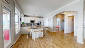 A spacious kitchen with white cabinets, a central island with stools, and a view through large windows.
