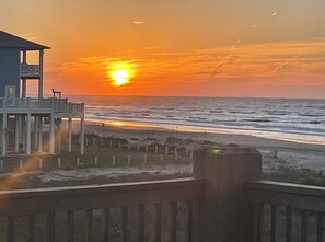 Vista a la playa con un atardecer, un muelle de madera y un edificio con balcón.