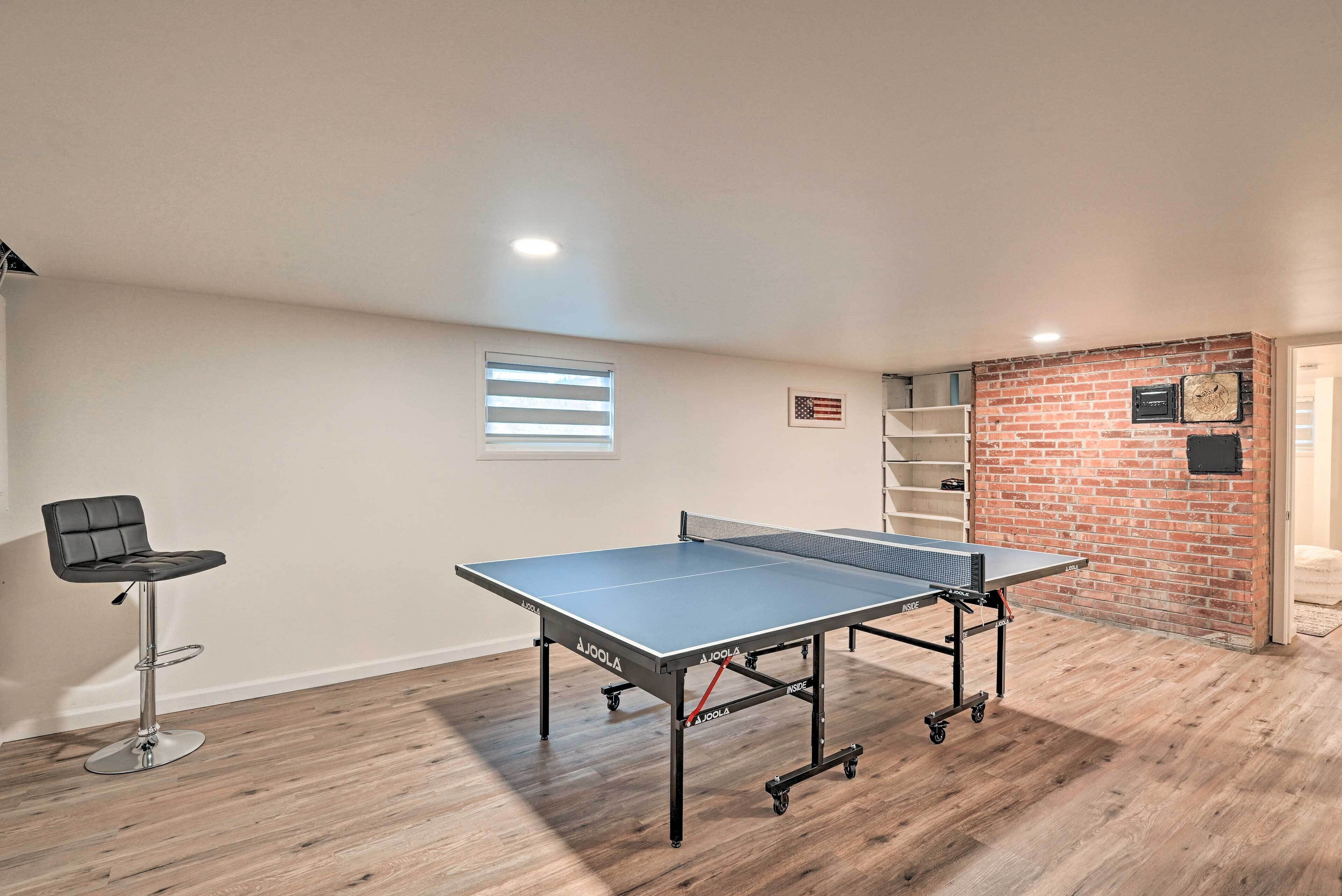 Une table de ping-pong dans une pièce avec un mur en briques, une fenêtre avec des stores et un tabouret de bar.