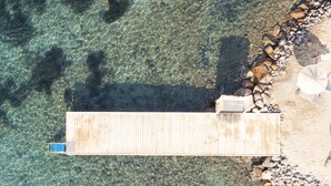 Vista aérea de un muelle que se adentra en aguas cristalinas, rodeado de rocas y una zona arenosa con una pequeña construcción.
