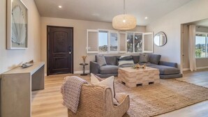 A spacious living room with a grey sofa, a coffee table, and a woven chair. There is a large window with shutters, a dark wooden door, and a hanging light fixture. - @Marbella Lane -  Charming Tranquil Hideaway (Yucca Valley)