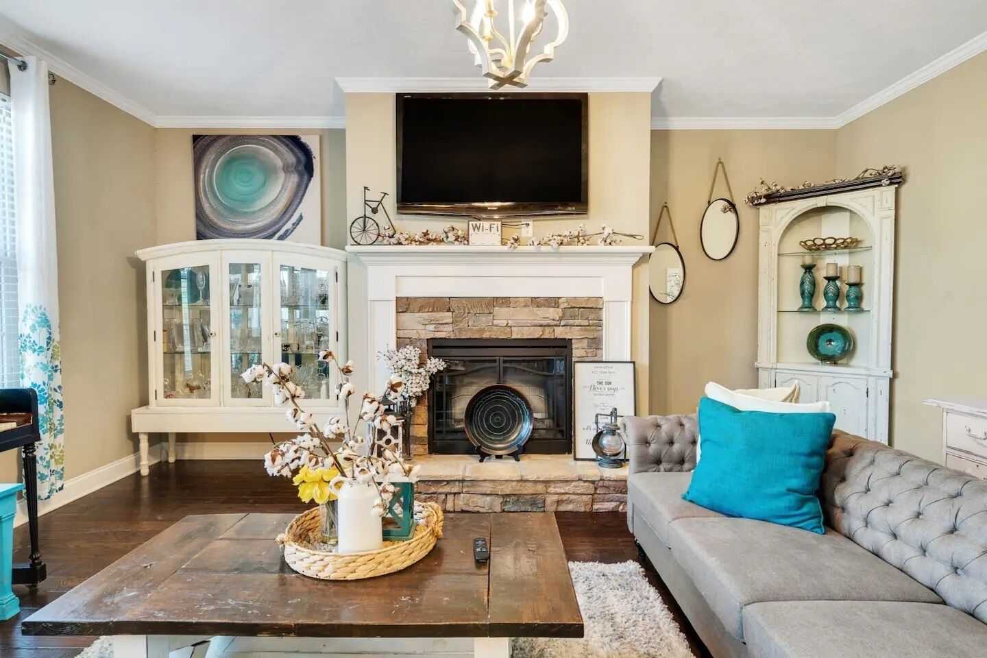 A living room with a fireplace, a television mounted above it, a cabinet with glass doors, and a sofa with a blue pillow.
