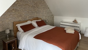 Une chambre avec un mur décoré d’un motif en pierre, une tête de lit en bois, un lit avec une literie blanche et marron, une table de chevet avec une lampe et une commode blanche avec des serviettes pliées.