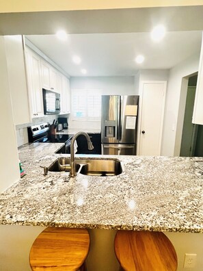 Una cocina con gabinetes blancos, electrodomésticos de acero inoxidable, encimera de granito y dos taburetes de madera.