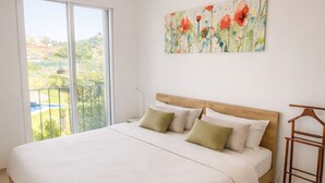 A spacious bedroom with a large bed, a wooden headboard, a hanging light fixture, a view of greenery through a large window, and a painting on the wall.