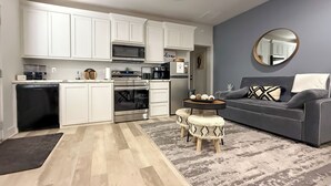 A modern kitchen with white cabinets, a microwave, oven, and coffee maker. There is a gray sofa with patterned cushions and a round mirror on the wall. - Relaxing Lubbock Studio ~ 6 Mi to Texas Tech (Lubbock)