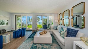 Una sala de estar moderna con un sofá blanco, sillones azules, una mesa de centro y grandes ventanales con vistas a zonas verdes y edificios.
