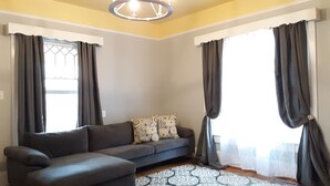 A living room with a grey sectional sofa, patterned cushions, a patterned rug, and large windows with dark curtains. - The Cam Grady, Restored, roomy, eclectic cottage in heart of Downtown Morganton (Morganton)