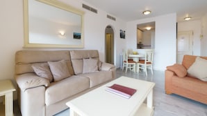 Un salon avec un canapé beige, une table basse blanche et un petit canapé orange. On aperçoit à l’arrière-plan un coin repas avec une table et des chaises.