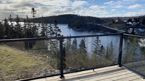 A wooden deck with a glass railing overlooking a lake, surrounded by trees and a few buildings in the distance. - Modern three bedroom with Panoramic Oceanview  (Hubbards)