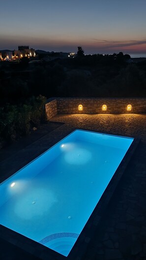 A rectangular swimming pool with a clear blue water surface, surrounded by stone walls and illuminated by wall-mounted lights.