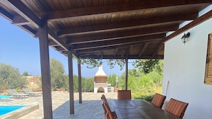 Un espace extérieur abrité doté d’un sol en pierre, d’une table en bois avec des chaises, et offrant une vue sur une piscine et sur la verdure.