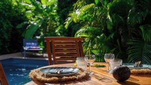 Une table de jardin, dressée avec des assiettes, des couverts et des verres, entourée d’une végétation luxuriante et d’une piscine.