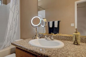A bathroom with a granite countertop, a white sink, a mirror with lights, a towel rack, and a bottle of perfume.