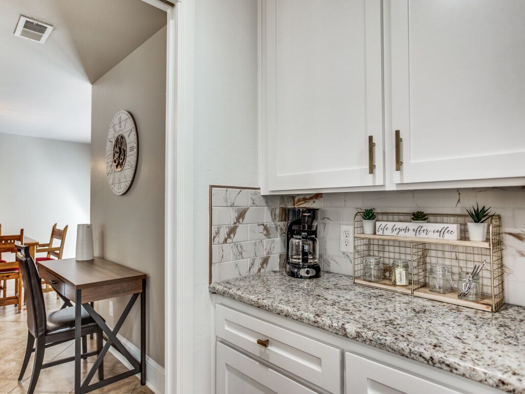 A kitchen with white cabinets, a granite countertop, a coffee maker, and a shelf with plants and jars.