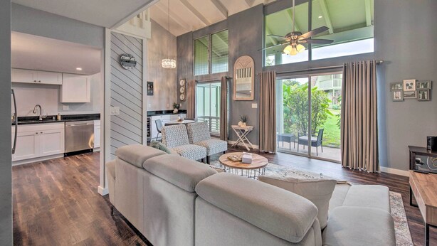 Una sala de estar moderna con un sofá grande, una mesa de centro y un comedor con mesa y sillas. Se observa una cocina con gabinetes blancos y un lavavajillas. La habitación tiene ventanas amplias con cortinas y vistas al exterior.