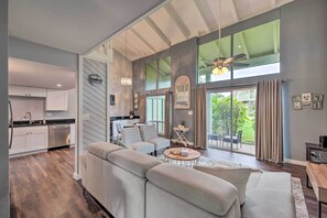 A modern living room with a large sofa, a coffee table, and a dining area with a table and chairs. There is a kitchen with white cabinets and a dishwasher. The room has large windows with curtains and a view of the outdoors. - ‘Coastal Escape’ Condo w/ Pool, Near Beaches! (Princeville)