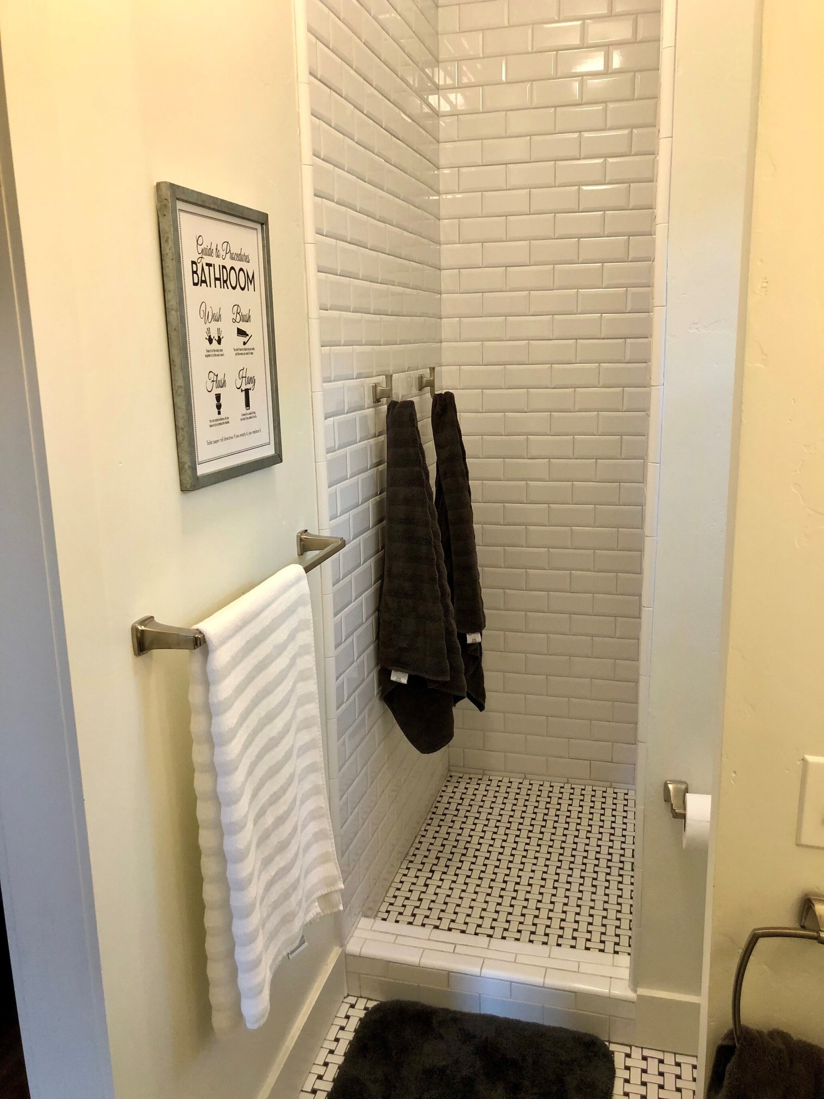 Een badkamer met witte wandtegels in metro-stijl, een douche met een vloer in zwart-wit patroon, een ingelijst overzicht van badkamerrituelen en twee handdoeken op een rek.
