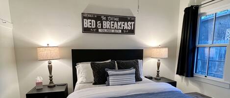 A bedroom with a bed, two bedside tables, a ceiling fan, a window with curtains, and a wall sign.
