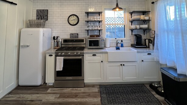 A kitchen with white cabinets, a stainless steel refrigerator, a microwave, and a stove. There is a clock on the wall and a chandelier hanging from the ceiling. - Downtown Cozy Cottage, two blocks from downtown and new! (Paso Robles)