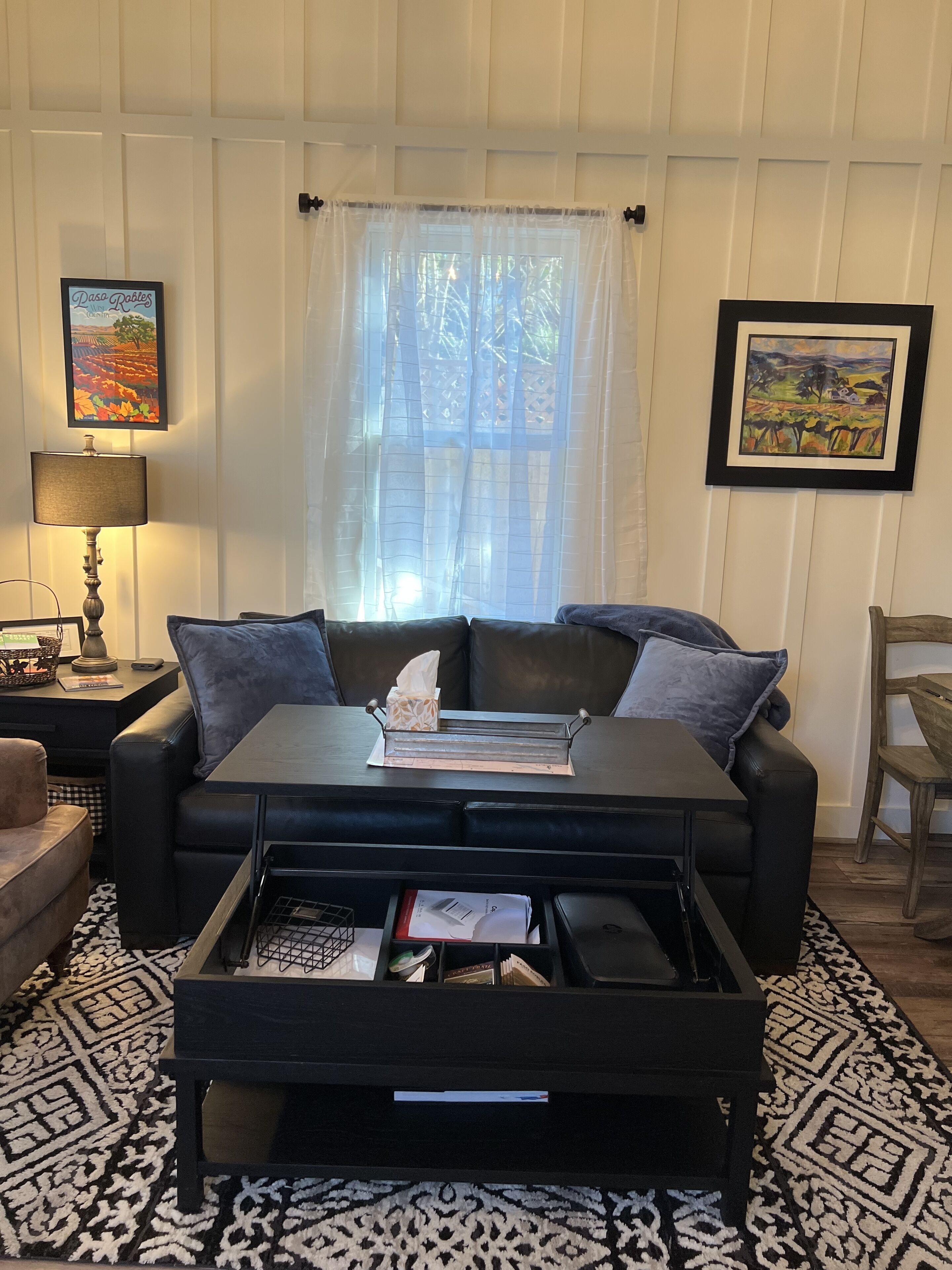 A living room with a black sofa, a coffee table, a patterned rug, and a window with sheer curtains.