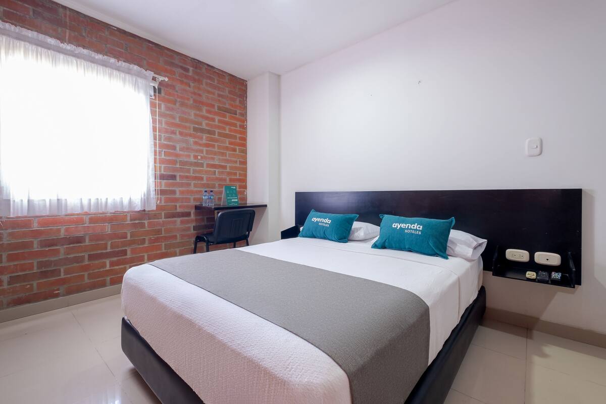 A hotel room with a brick wall, a bed with a grey and white bedspread, two blue pillows with the hotel name, a desk with a chair, and a window with white curtains.