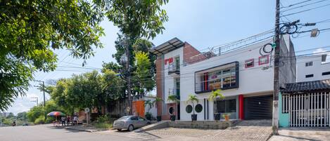 A street view with a modern building featuring a balcony, a parked car, and a sidewalk with trees.