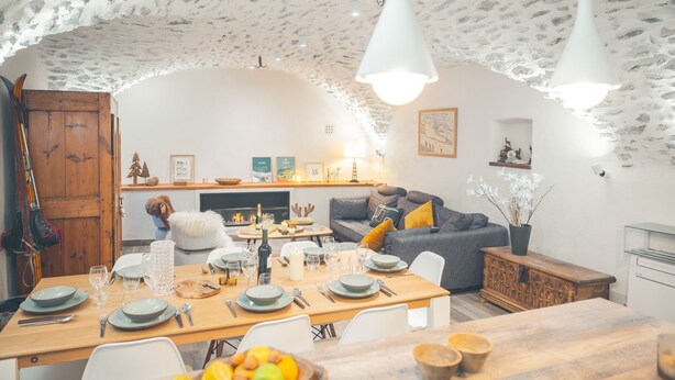 Un coin repas chaleureux avec une table en bois dressée pour quatre personnes, une cheminée et un canapé agrémenté de coussins.
