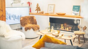 Un salon chaleureux avec une cheminée, un fauteuil blanc, un coussin jaune et une petite table en bois sur laquelle sont posés des verres et une bougie.