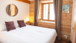 Une chambre avec un mur en bois, un lit avec une literie blanche, deux coussins à motifs, une table de chevet avec une lampe, une fenêtre avec des rideaux et un miroir fixé au mur.