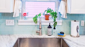 Una cocina con un lavamanos de acero inoxidable, una ventana con cortinas blancas y una planta en maceta en el alféizar.