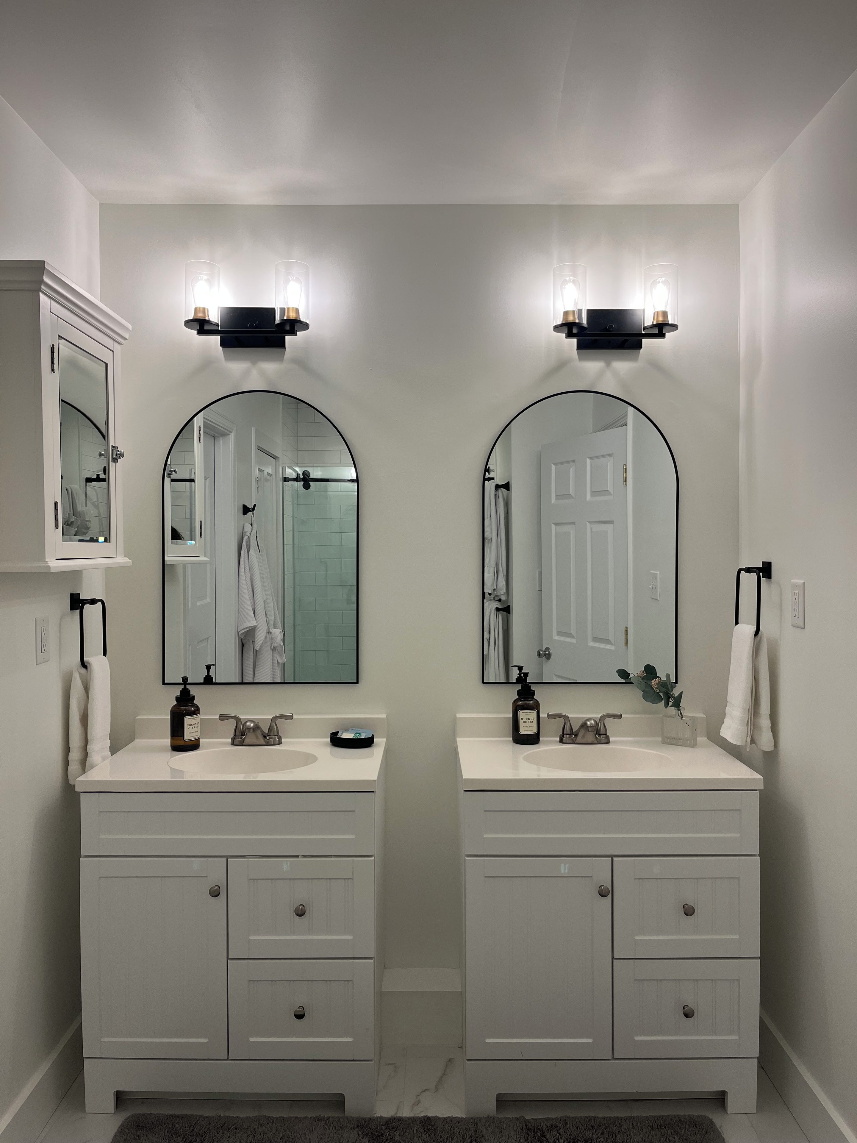 Une salle de bain avec deux lavabos, chacun étant surmonté d’un miroir. On trouve des placards sous les lavabos, et un porte-serviettes est visible sur le mur.