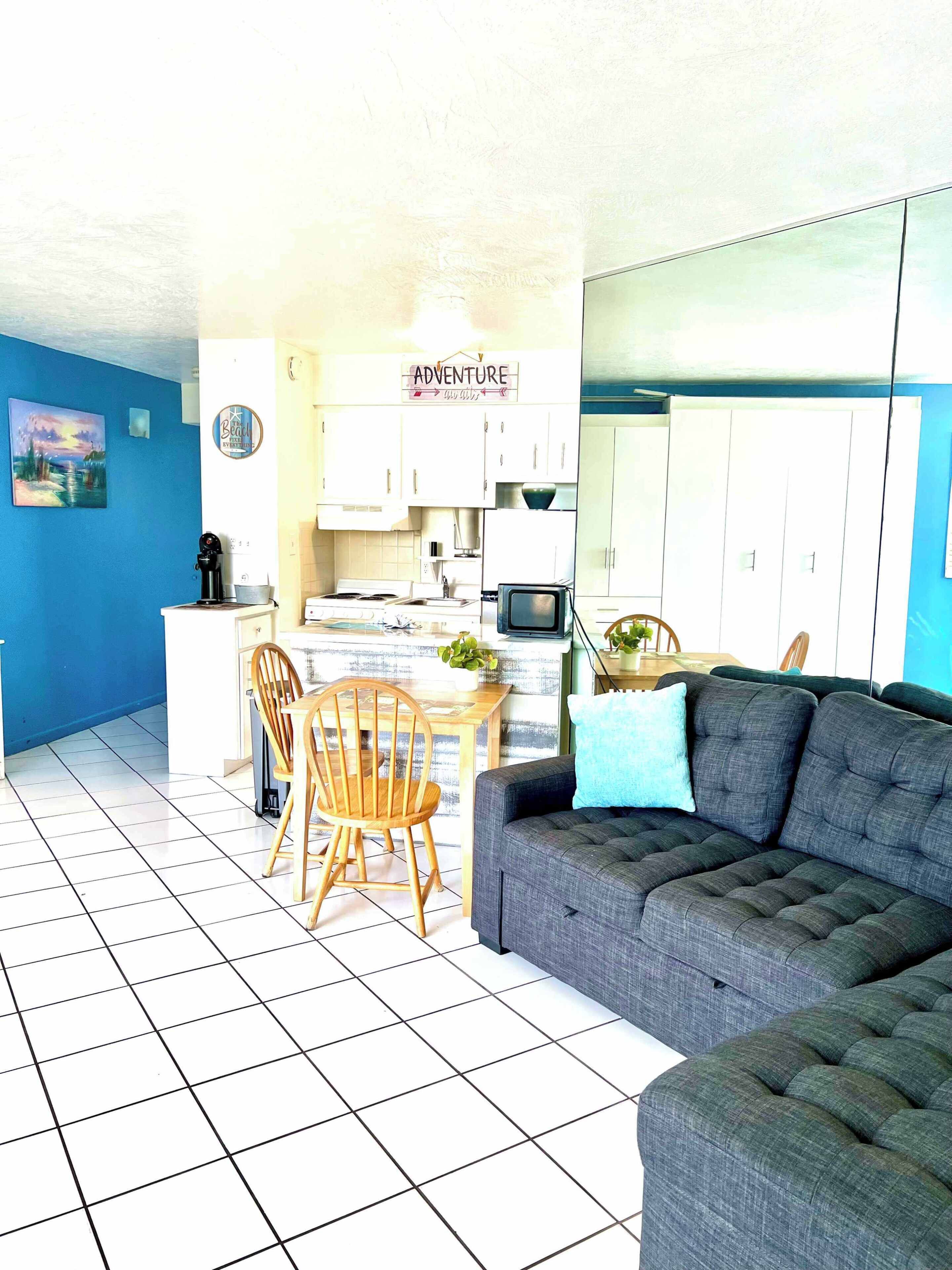 Un salon avec un canapé gris, une table à manger en bois avec des chaises, un coin cuisine avec des placards et un four à micro-ondes, et un mur bleu.