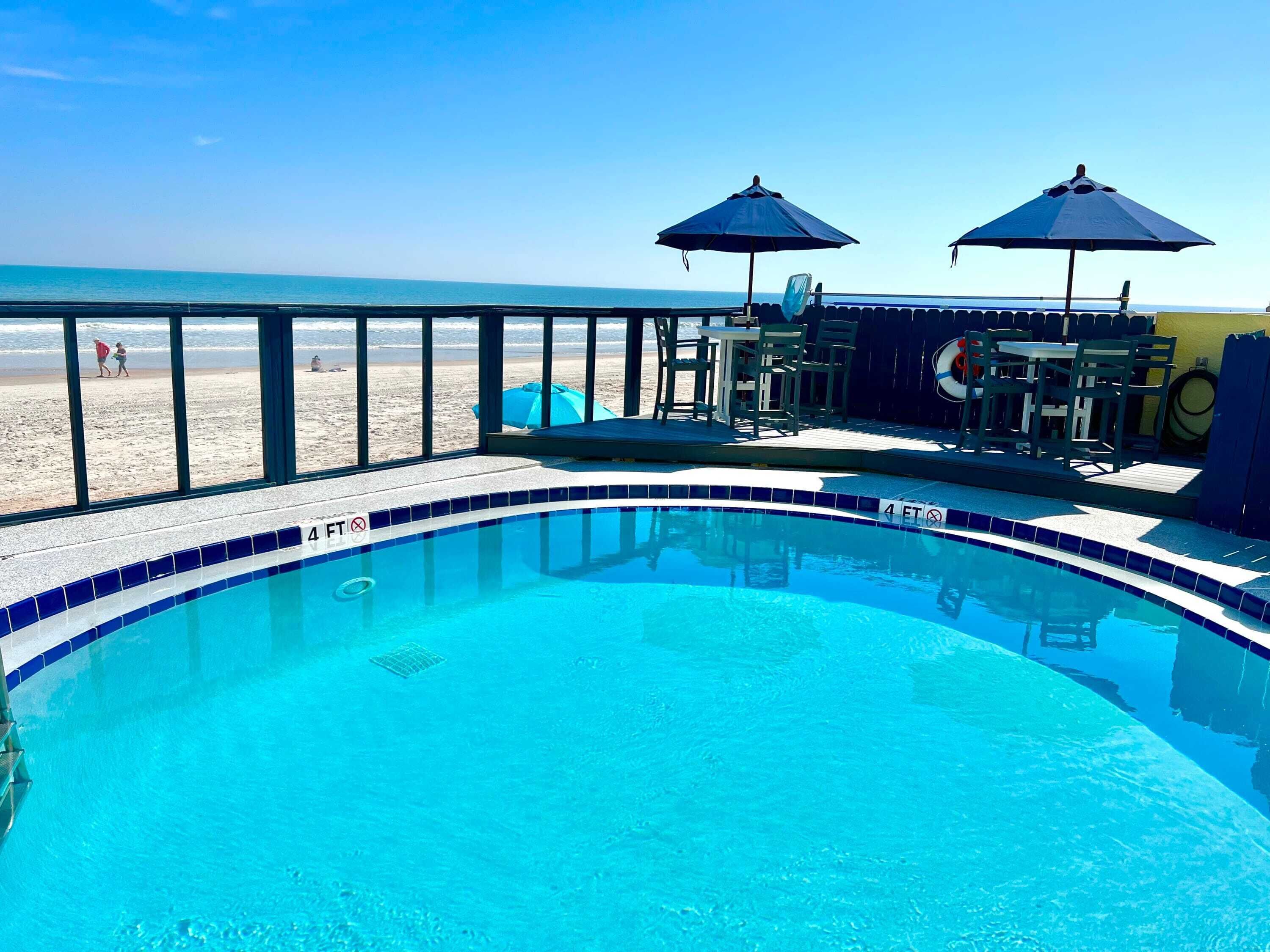Ein Schwimmbecken mit einer 1,2-Meter-Tiefenmarkierung, Sitzgelegenheiten im Freien und Blick auf den Strand.