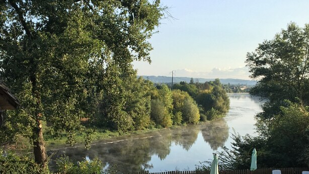 Une terrasse au bord de la rivière, avec une table en bois et des chaises longues blanches, entourée d’une clôture en bois et d’une végétation luxuriante.