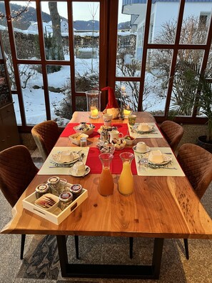 A dining table set with various dishes, glasses, and a gnome decoration, with a snowy landscape visible through large windows. - Pension Lambrecht (Sankt Lambrecht)