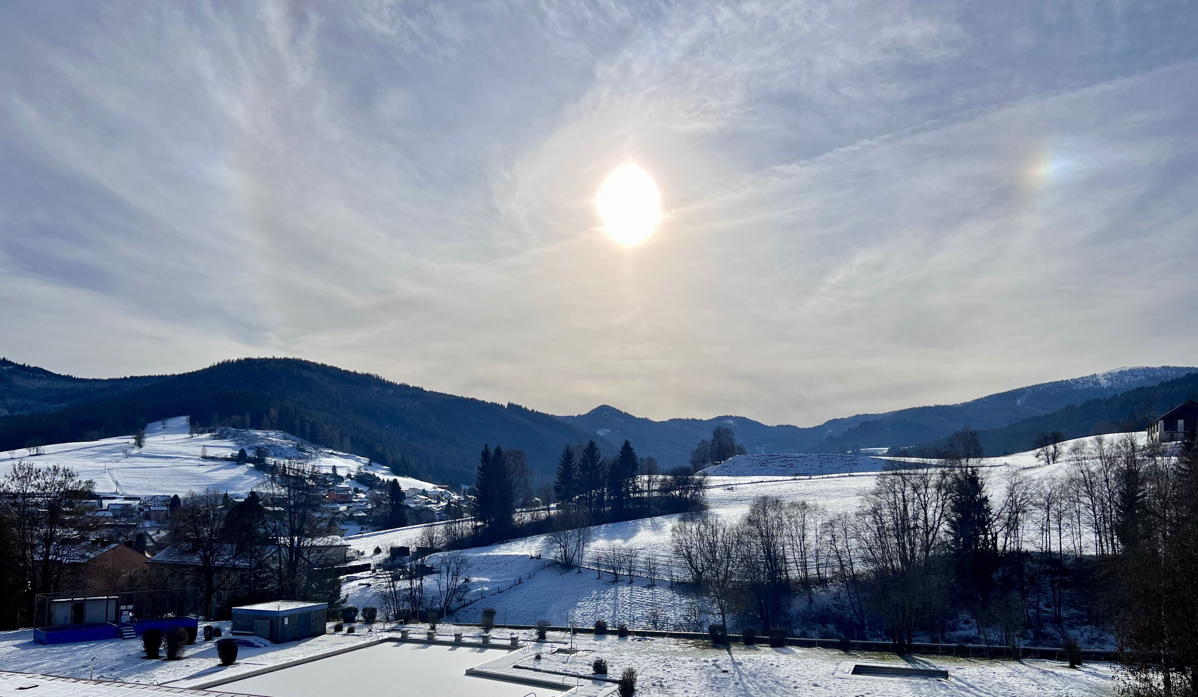 A snowy landscape with a clear sky, a sun shining, and a village with buildings and trees.