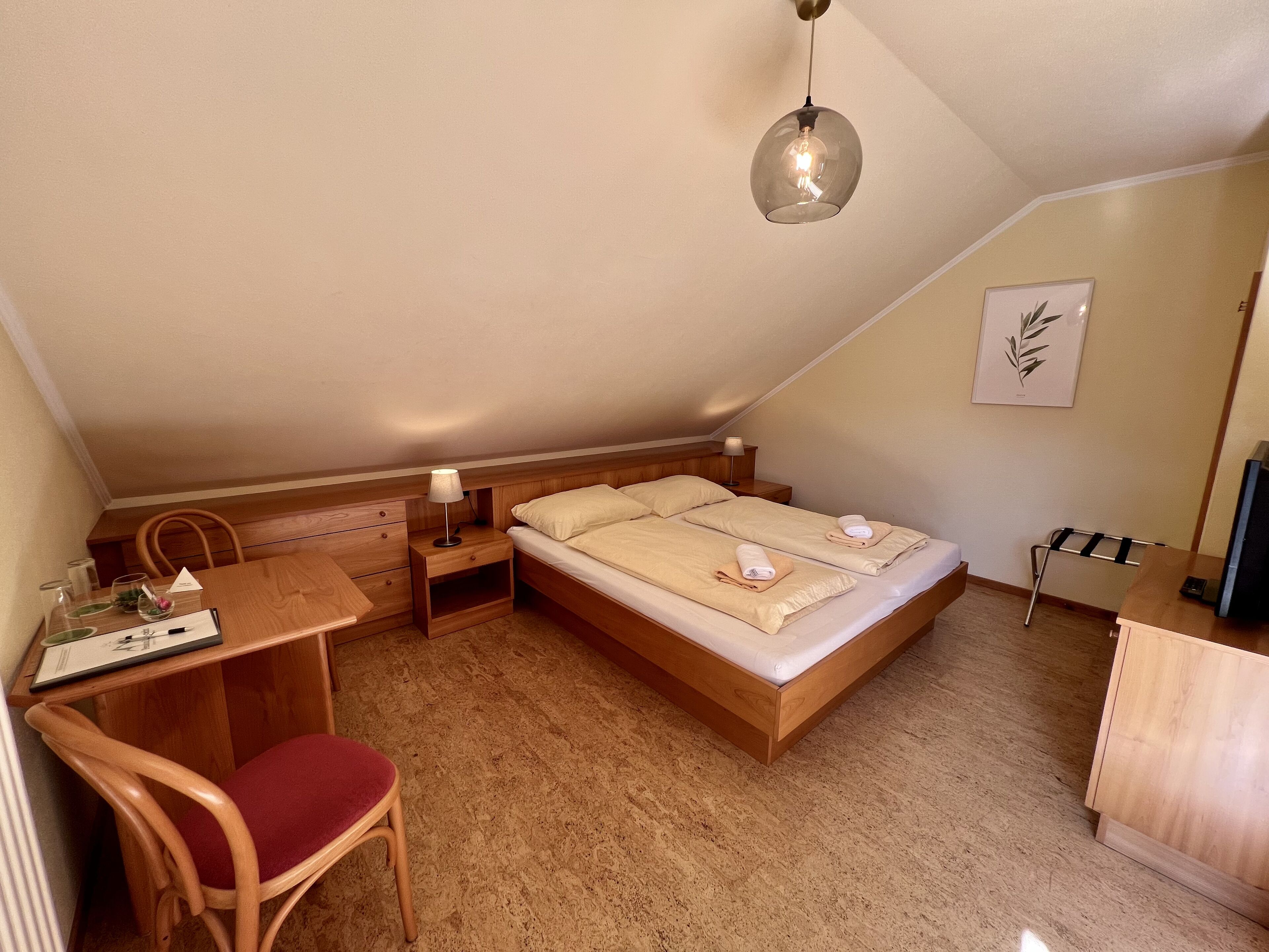 Une pièce chaleureuse et bien éclairée, avec un bureau en bois, une chaise, un lit dont la literie est soigneusement disposée, et un luminaire suspendu.
