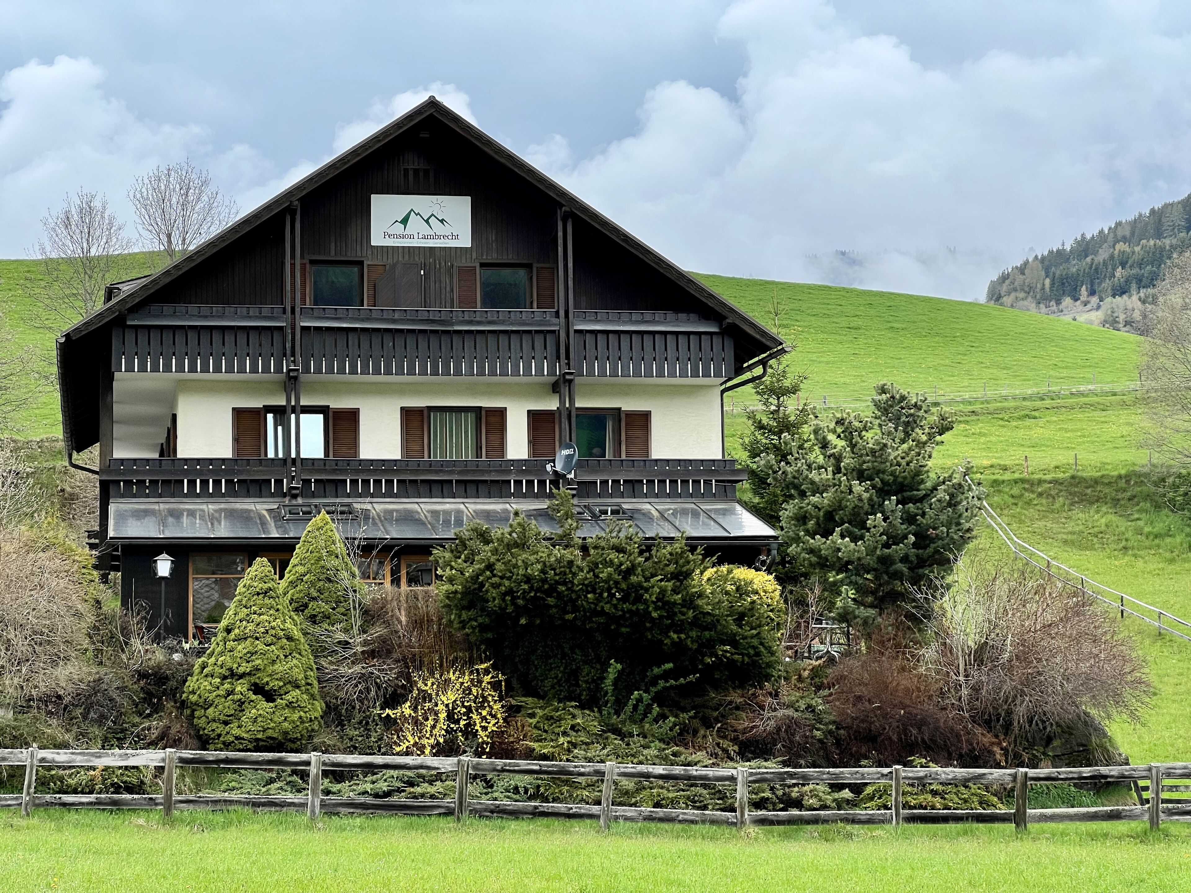 A two-story wooden building with a balcony, a sign reading "Pension Lembrecht," and surrounded by greenery.