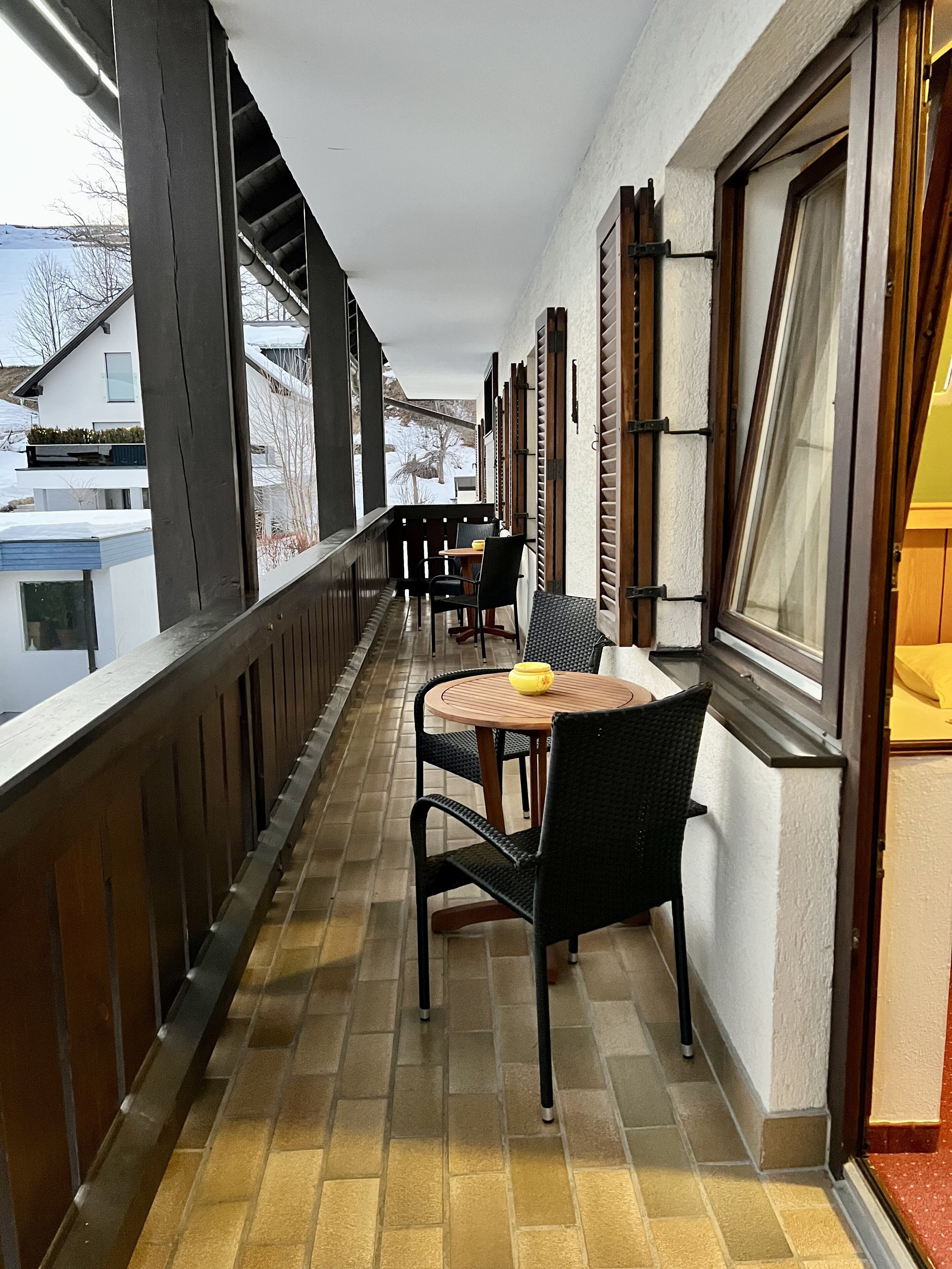A balcony with a table and chairs, a window with shutters, and a view of a snowy landscape.