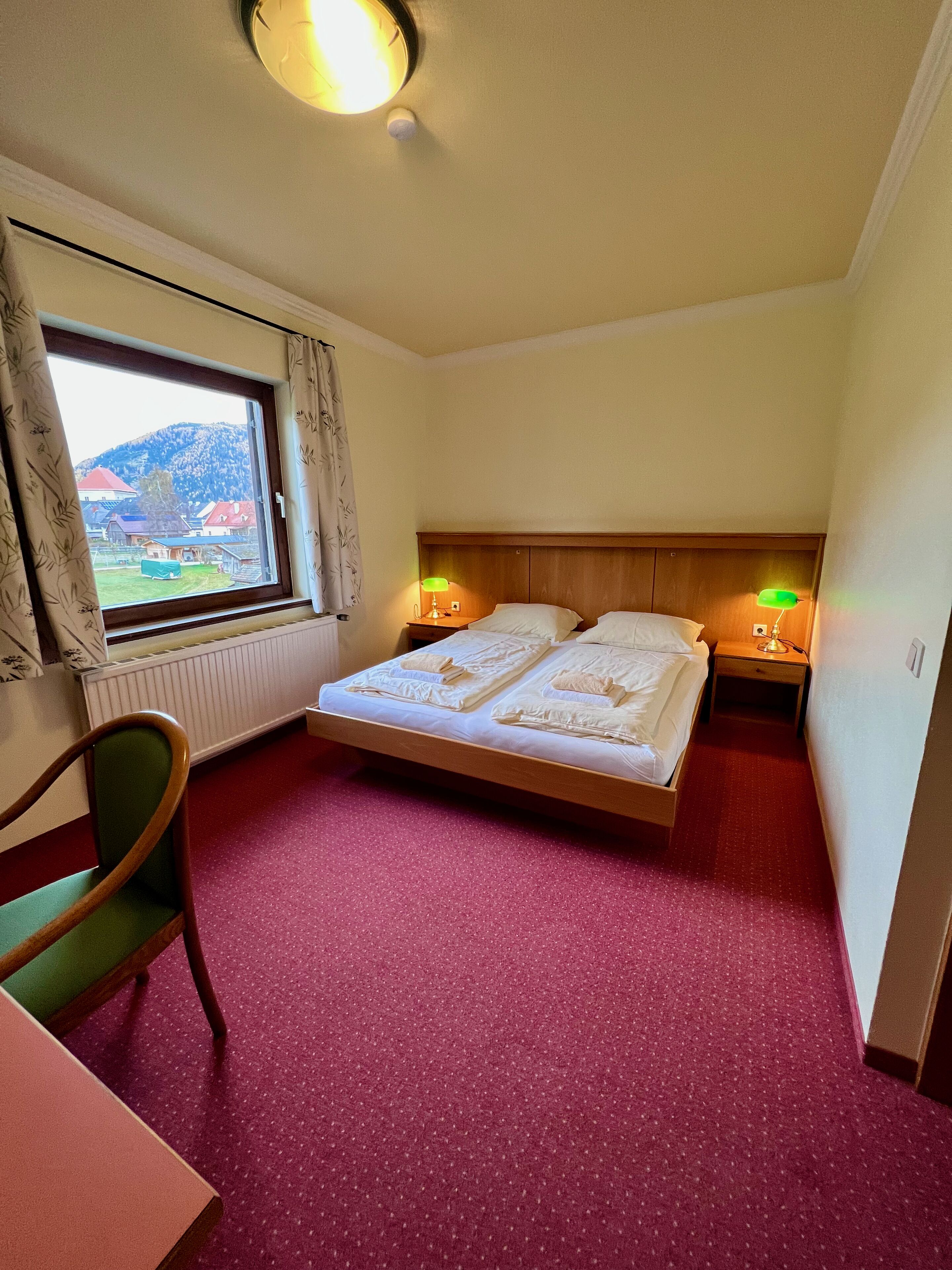 Habitación de hotel con cama, dos mesitas de noche con lámparas, una silla, ventana con cortinas y un radiador.