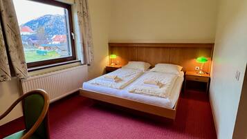 Habitación de hotel con cama, dos mesitas de noche con lámparas, una silla, ventana con cortinas y un radiador.