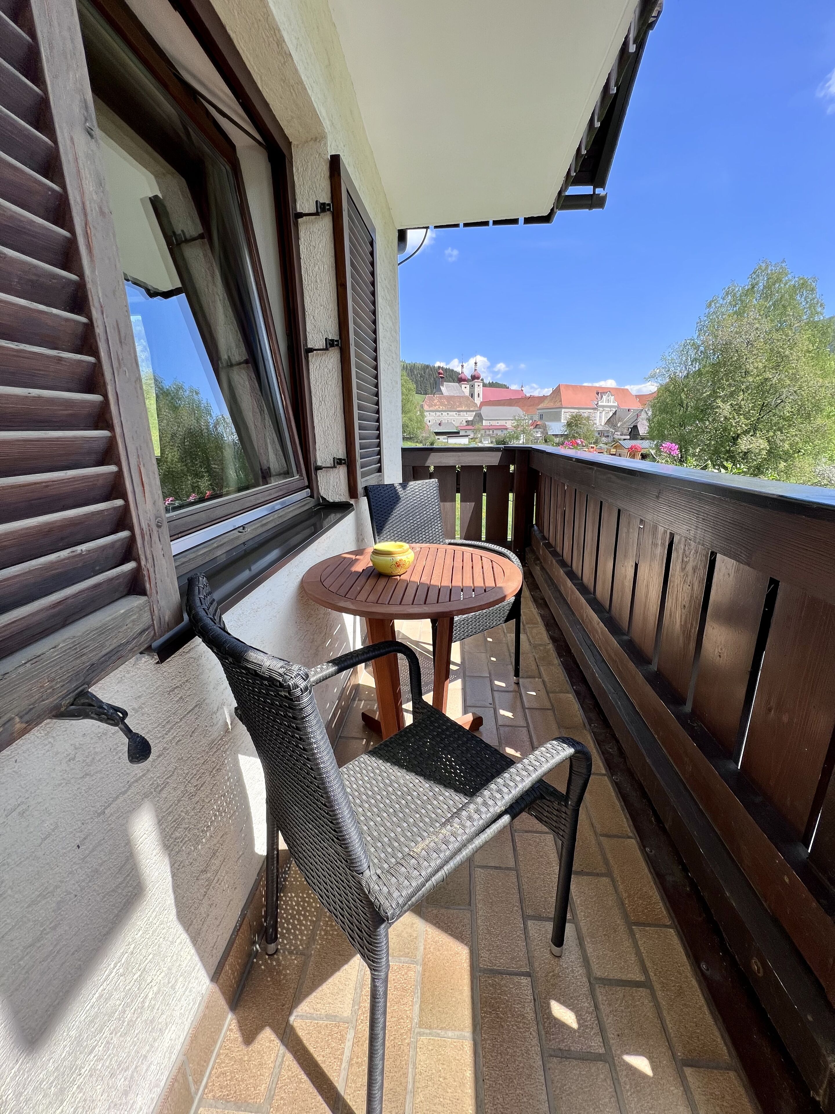 Un balcón con mesa y sillas, una ventana con persianas, y vistas a edificios y árboles.