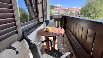 Un balcón con mesa y sillas, una ventana con persianas, y vistas a edificios y árboles.