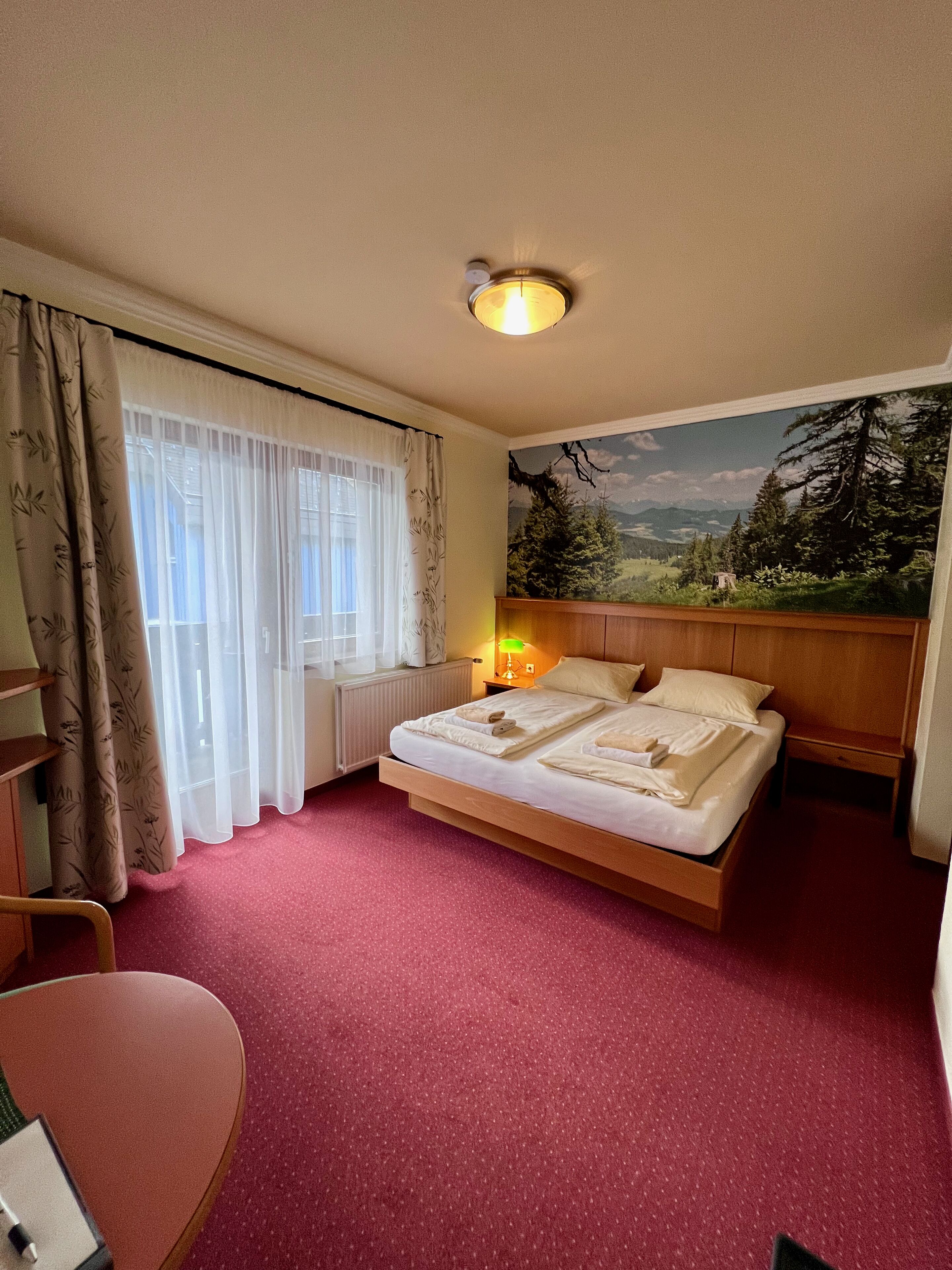 Ein Hotelzimmer mit Bett, Nachttisch, Lampe, einem Fenster mit Vorhängen, einem Sessel und einem Bild an der Wand.