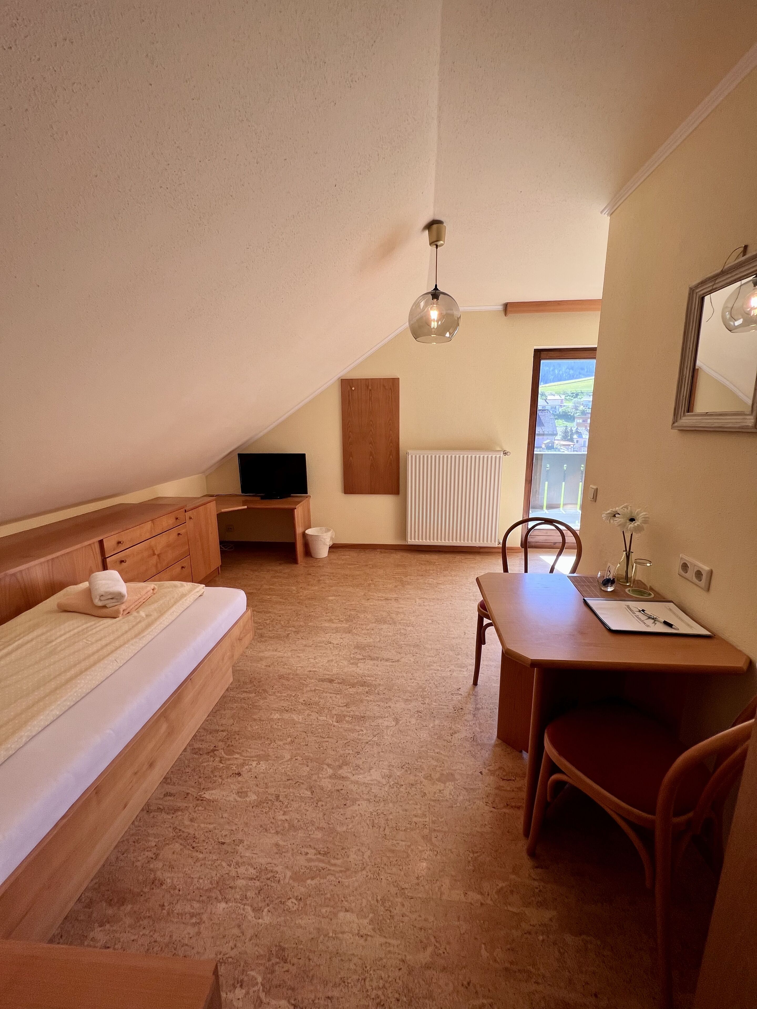 A small, cozy room with a bed, a desk, a chair, a television, and a window with a view.
