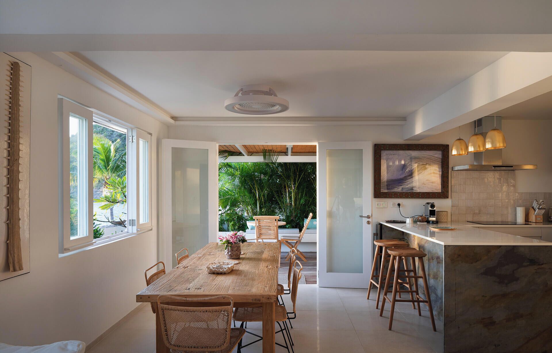 Une cuisine moderne dotée d’un îlot central, d’un coin repas avec une table et des chaises en bois, et offrant une vue sur un jardin luxuriant grâce à une porte vitrée.
