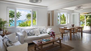 Un'ampia zona giorno con un divano bianco, un tavolino in legno e un tavolo da pranzo per quattro persone. Grandi finestre offrono vista sull'oceano e sulle palme.