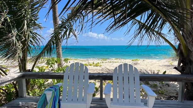 Two white Adirondack chairs on a wooden deck overlooking a beach with clear turquoise water and palm trees.